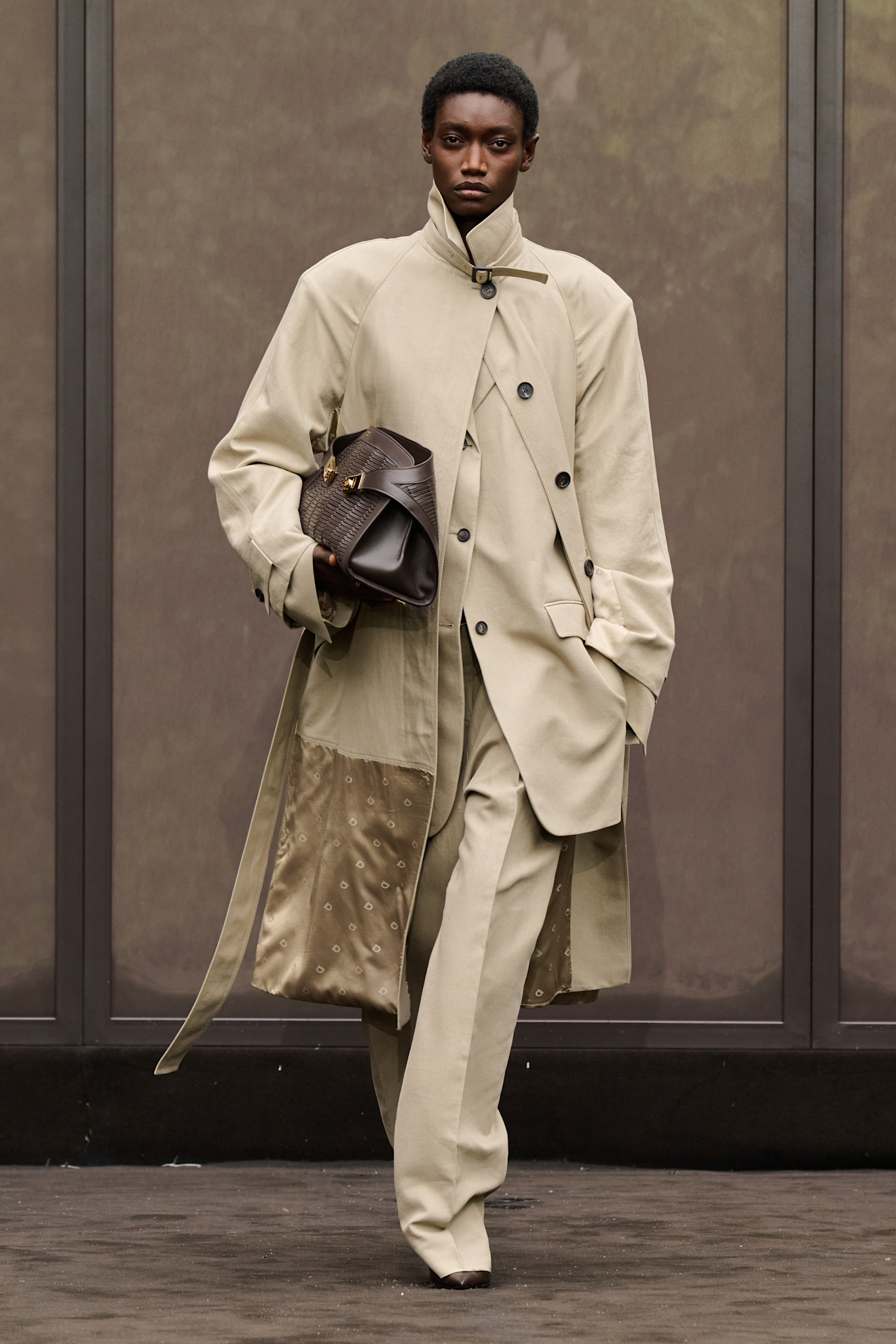 Layering staples for spring 2026 are shown in a photo from Ferragamo's runway show, with a model walking while wearing a long tan trench coat with a funnel neck layered on top of a matching tan blazer, and tan pleated trousers styled with a dark brown snakeskin clutch bag and brown pointed heels