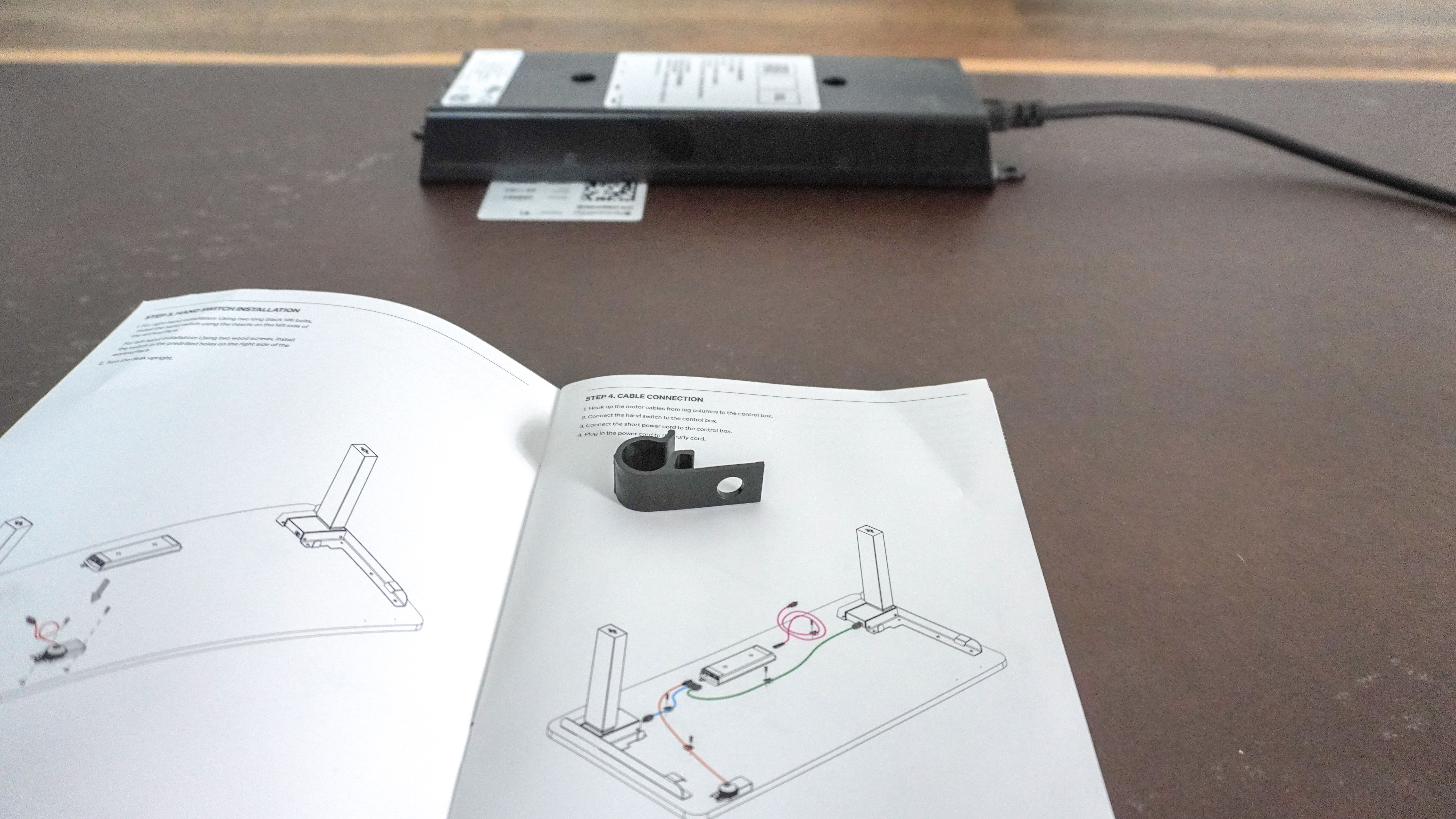 The instruction manual for the Herman Miller Coyl Gaming Desk with a plastic cable management hook on top of it with the desk's control box in the background