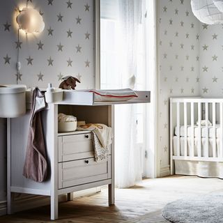 White changing table with drawers, grey wall and white cot with grey rug and starry wallpaper