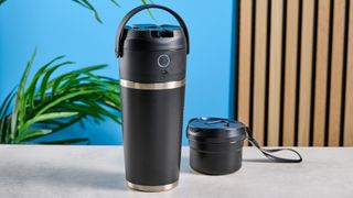 the Nutribullet Flip with a black insulated tumbler and a portable battery-powered blade photographed with a blue background. The tumbler has a carry strap and a sippy lid you can interchange, and an on/off button on the base.