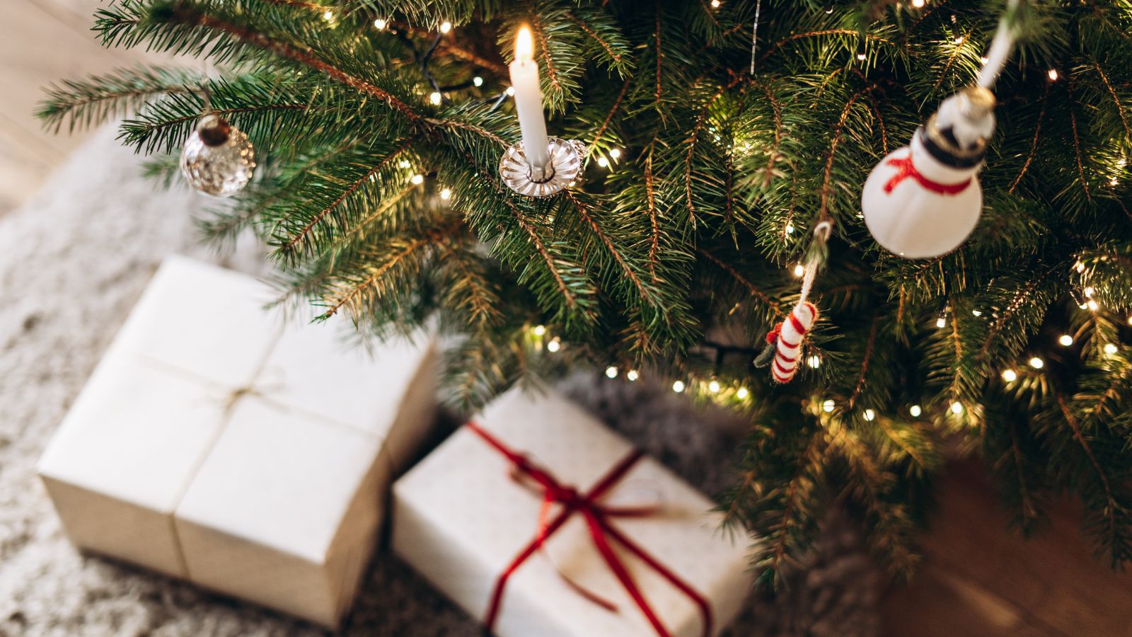 Festive Christmas tree decorated with white and silver ornaments, candles, and twinkling lights, standing in a cozy room. Christmas presents are under the tree. 