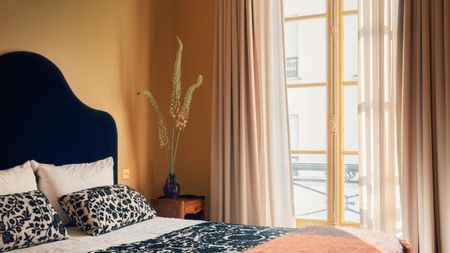 A bedroom with dark yellow walls, a beige ceiling, and French doors leading onto a balcony. Sheer beige curtains over the doors, and a bed with a black upholstered headboard, cream and black patterned cushions and a throw, and an orange-red blanket. 