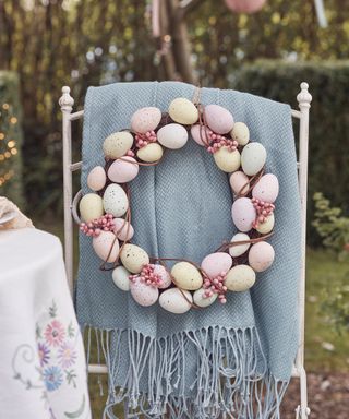 A pastel egg-colored garland on blue thrown on a white metal chair next to a white tablecloth in the garden