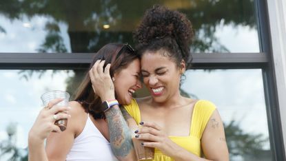 two women laugh while they hug each other outdoor near a building, wearing casual summer clothing