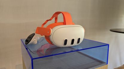 An orange Meta Quest 3 and its controllers on a blue plastic plinth in front of a blank wall