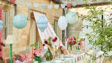 An outdoor dining scene with floral paper lanterns and bunting and a table with candles and plates, with a stone house in the background.