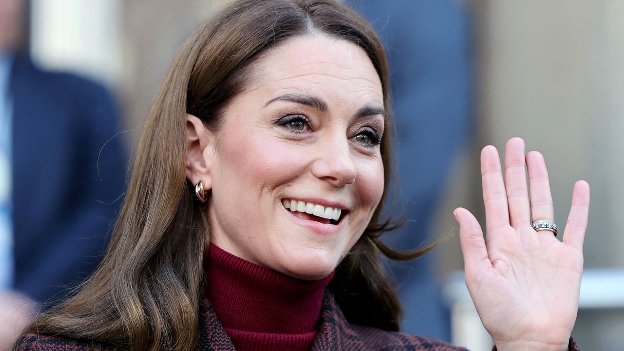 Kate Middleton smiling and waving wearing a red turtleneck and gold hoop earrings