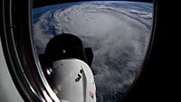 a hurricane is seen through the window of an orbiting spacecraft, with a white space capsule in the foreground