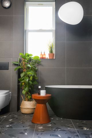 Grey bathroom with large format wall tiles, terrazzo floor tiles, black freestanding bath and terracotta side table