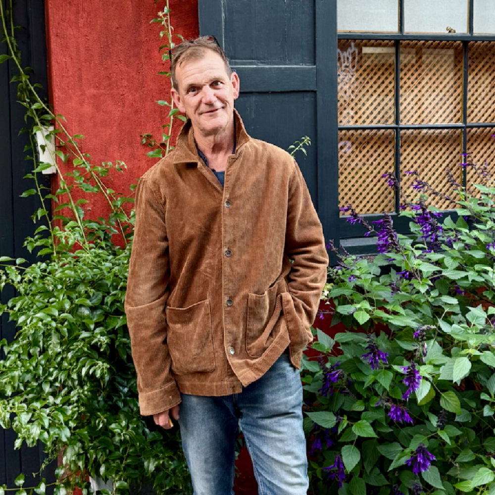 male wearing brown jacket and jeans stood in front of plants