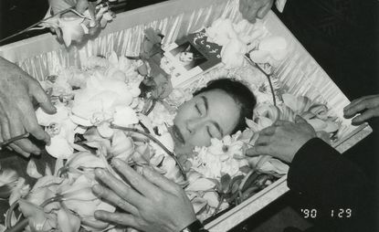 A black and white photograph of Nobuyoshi Araki’s deceased wife in a coffin with various hands reaching in to place flowers