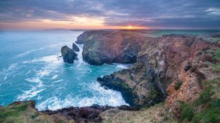 Sunrise over Hell's Mouth Cornwall, UK. Sea crashes against cliffs, with sun peeping over horizon beneath overcast clouds