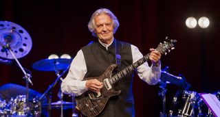 John McLaughlin wears a black waistcoat and white shirt as he plays his PRS signature guitar onstage.