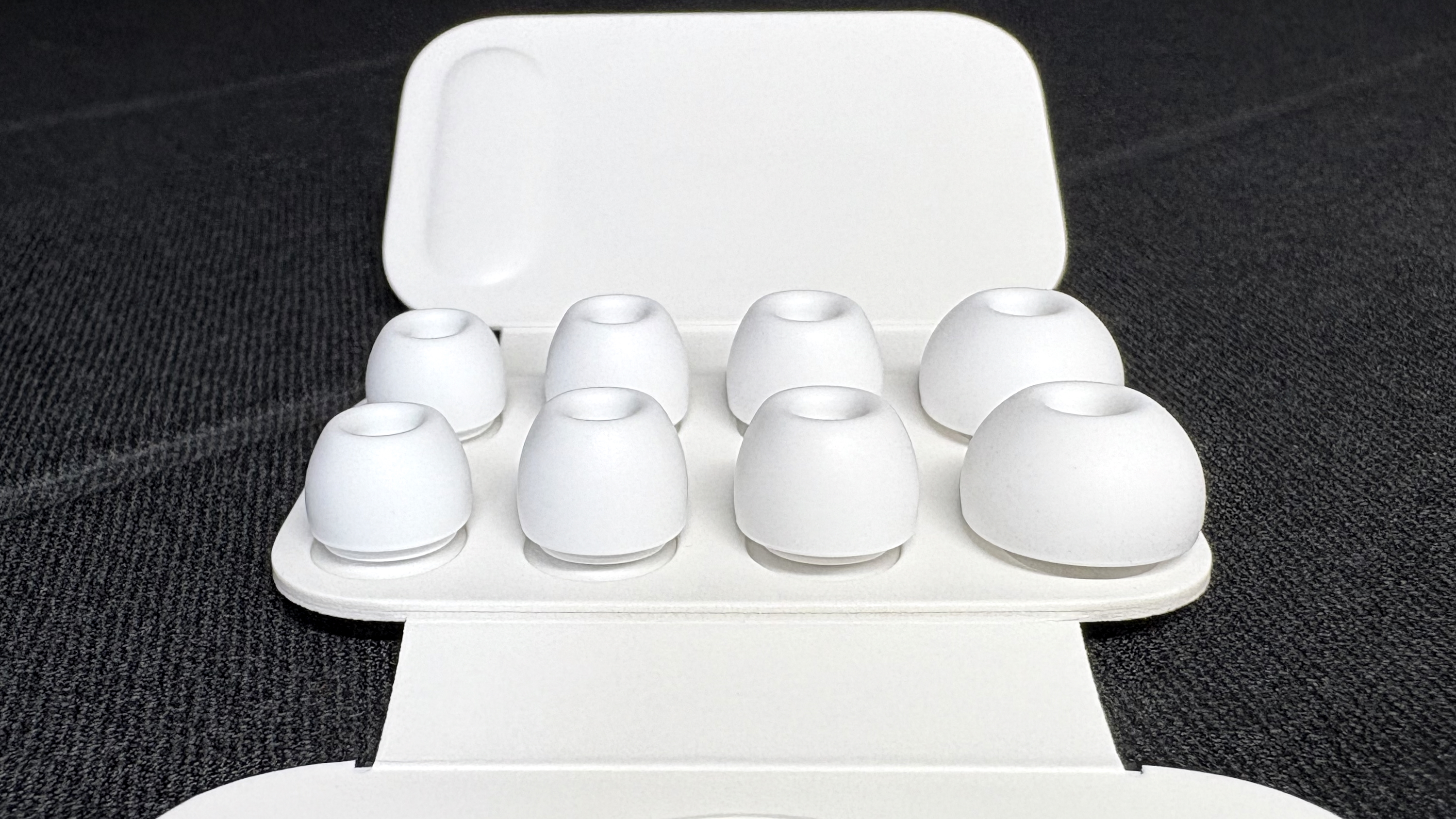 Apple AirPods Pro 3 either worn by a man standing in front of a red-brick wall, or on a table with gray twine cover
