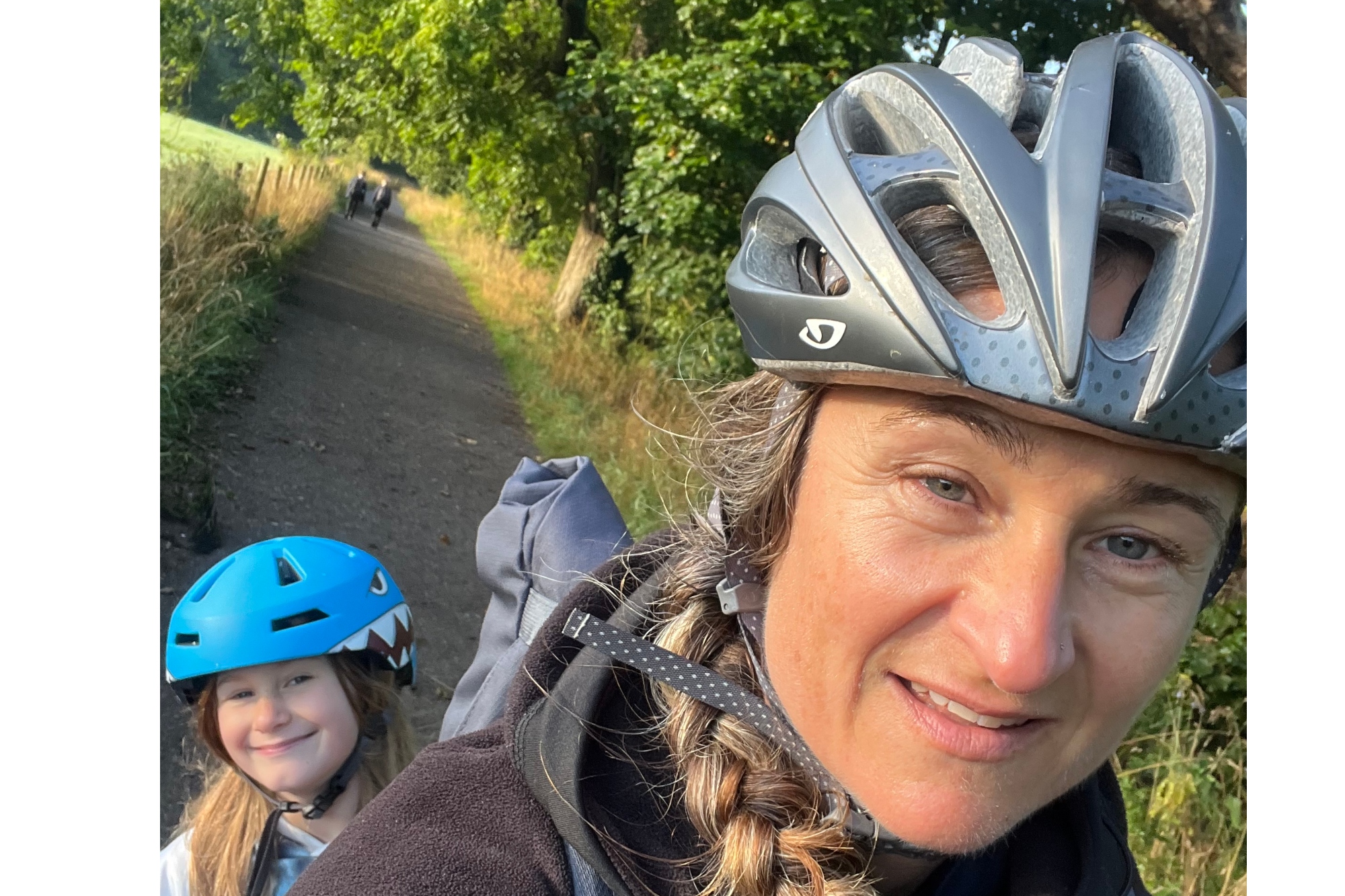 A woman is looking at the camara smiling with a helmet on. In the background is a child with a blue helmet. They are on a cargo bike