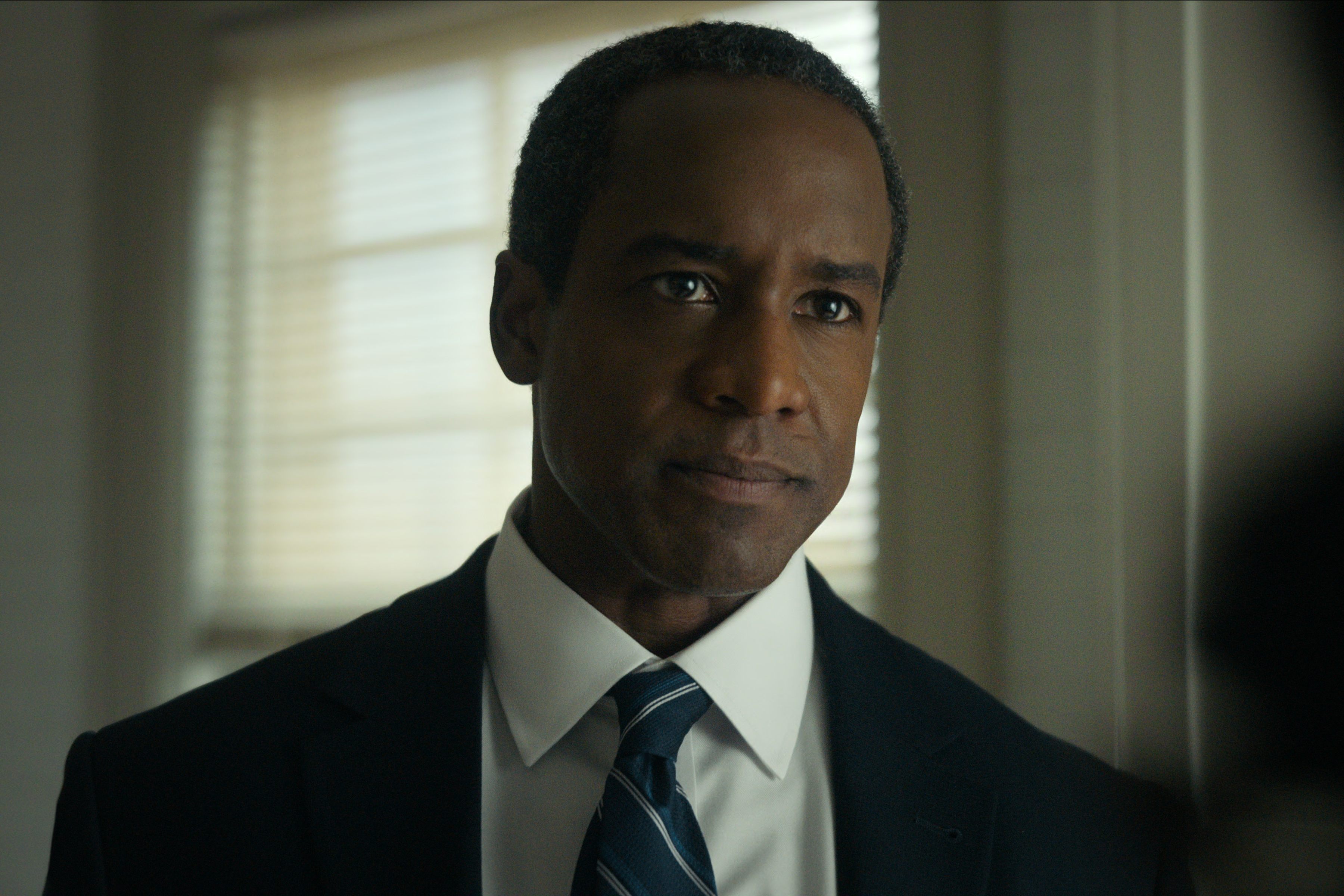 Albert Jones as Deputy Director Aiden Mosley wearing a suit and tie standing in front of a window looking stern in a still from the night agent season 3