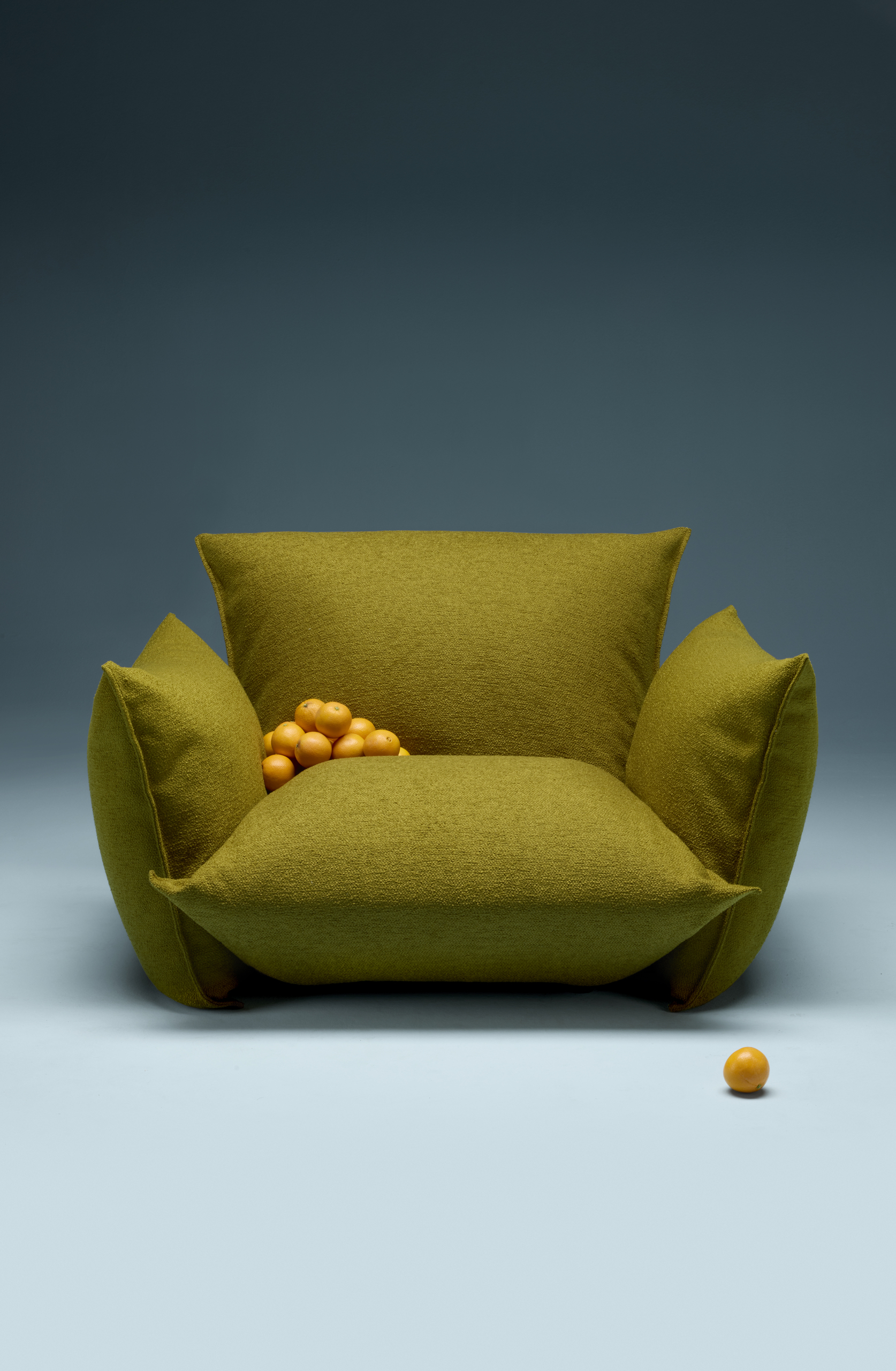 Image of an olive-green, contemporary-style, overstuffed armchair with oranges piled in the corner. The chair is in an all-blue showroom.