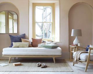 Cozy living room with daybed with light wooden frame, cream upholstered cushion, dressed with colorful cushions, window between two arch alcoves, pink painted walls, wooden flooring, textured natural rug