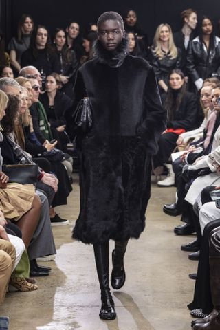 Proenza Schouler model walking in the F/W 24 show wearing a black fur funnel-neck coat with a black clutch bag and black boots.