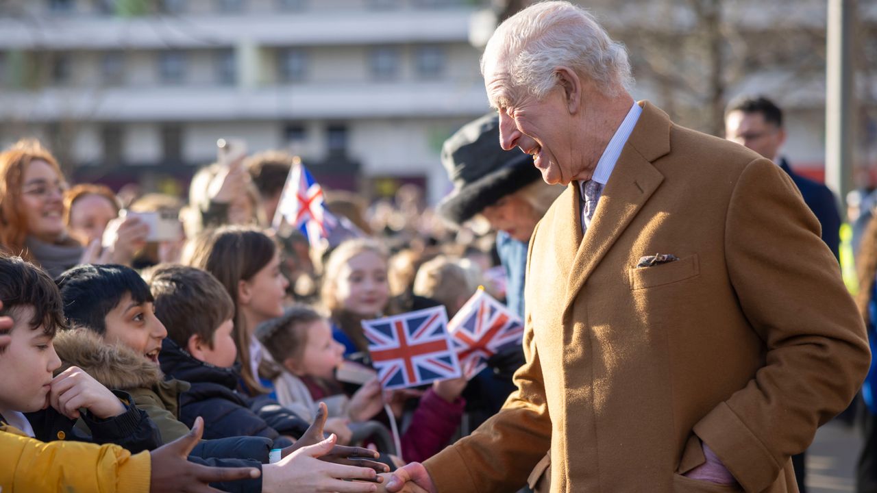 King Charles wearing a camel colored coat shaking hands with kids outside who are waving union jack flags