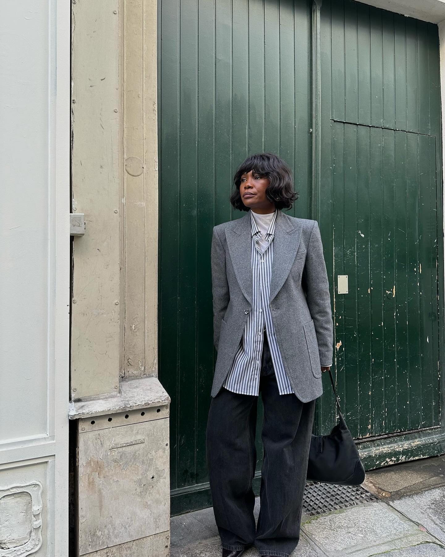 Fashion person Fran Fyne wears a grey blazer, striped shirt, white turtle neck and black baggy jeans. She is stood in front of a green door in a recent image.