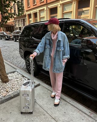 Varley's Lara Mead holding a suitcase handle and wearing a pink matching set with a denim jacket and red baseball cap.