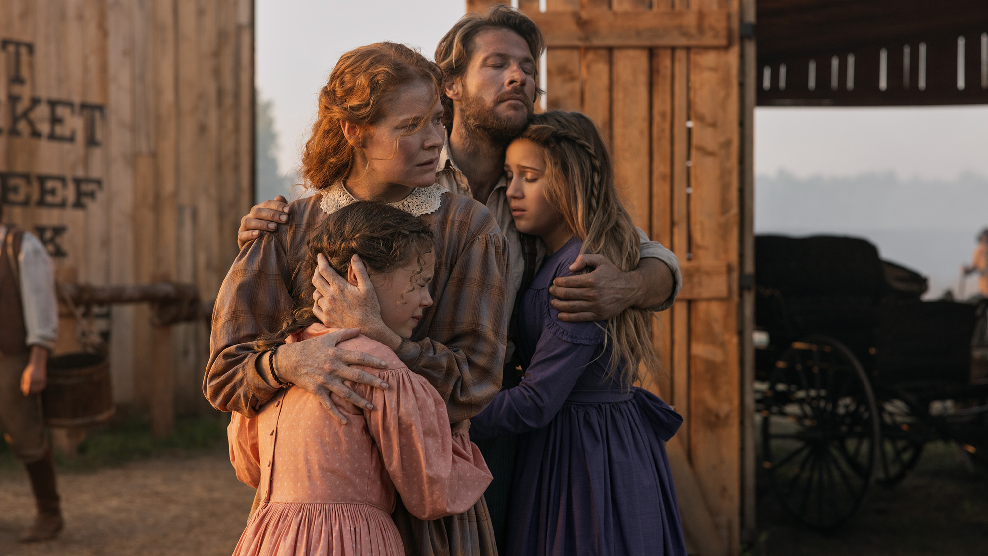 Luke Bracey and the main cast wear prairie cotton fits with wooden outbuildings in the background on Little House on the Prairie. 