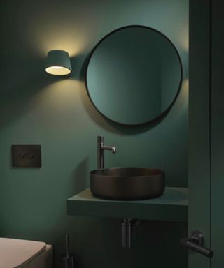 A dark green bathroom with a black rimmed mirror, black basin and back taps. A black wall light adds a gentle glow