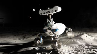 A close-up shot of a small, white and blue rover on the simulated lunar surface in a low-light environment. The rover has six wheels, a large sensor head on top, and a compact body.
