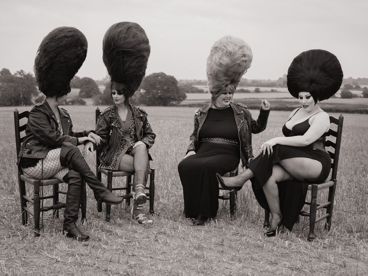 Black and white photo of four members of the Rebel Dykes in studded leather jackets, fishnets and towering beehive wigs, sitting on wooden chairs in a stubble f