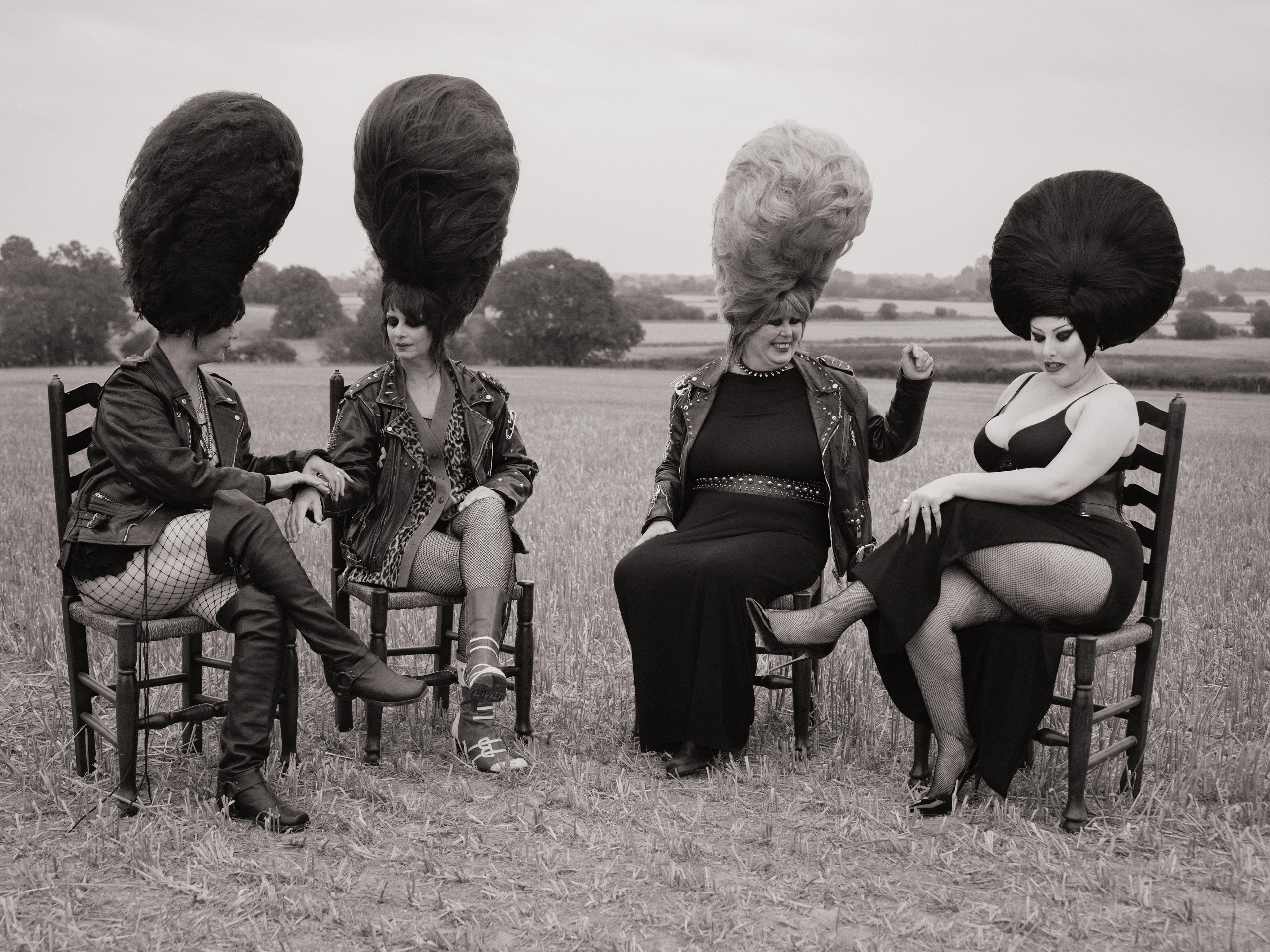 Black and white photo of four members of the Rebel Dykes in studded leather jackets, fishnets and towering beehive wigs, sitting on wooden chairs in a stubble field.
