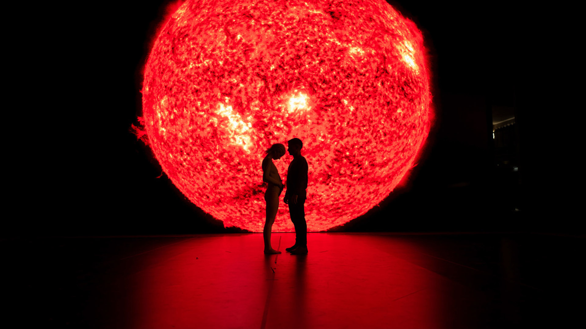 Two people stand silhouetted in front of a fake sun in the movie Ema