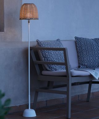 Chic outdoor floor lamp in white and woven wicker stood by chic sofa with patterned cushions.