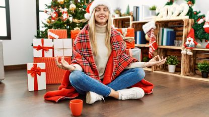 A woman wearing a Santa hat and wrapped in a Christmas blanket sits in front of a Christmas tree looking very relaxed.