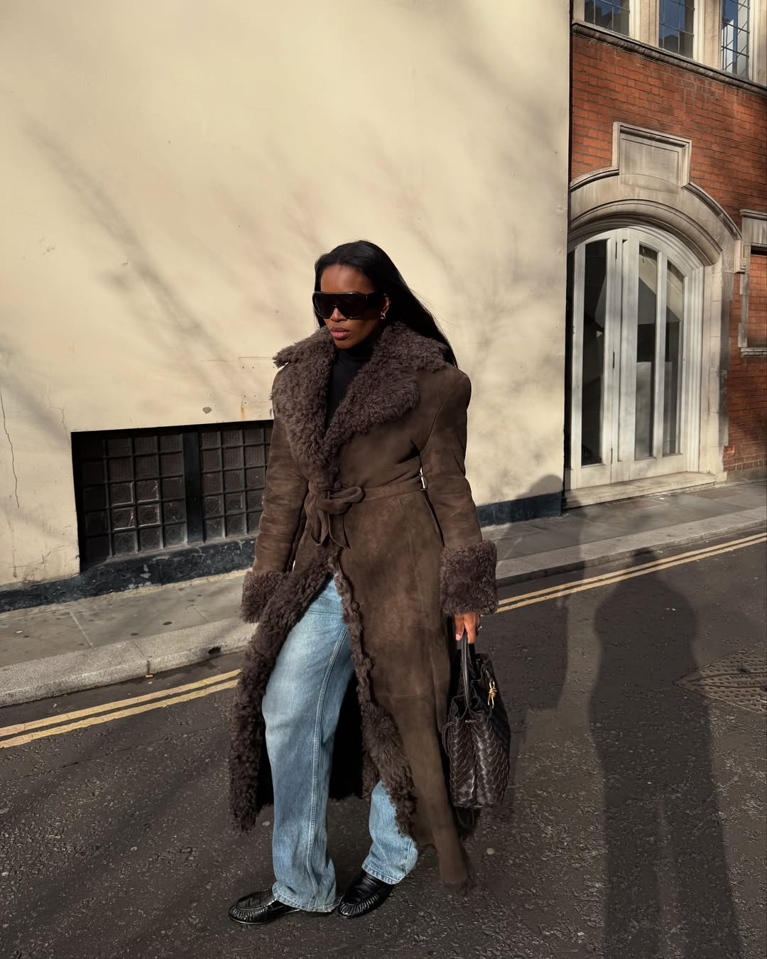 A woman wears gathered loafers with jeans and a brown coat.