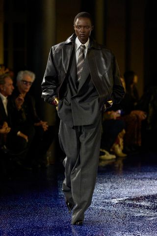 An S/S 25 model wearing a suit, tie, leather jacket, and gold cuff bracelets at the Saint Laurent show.