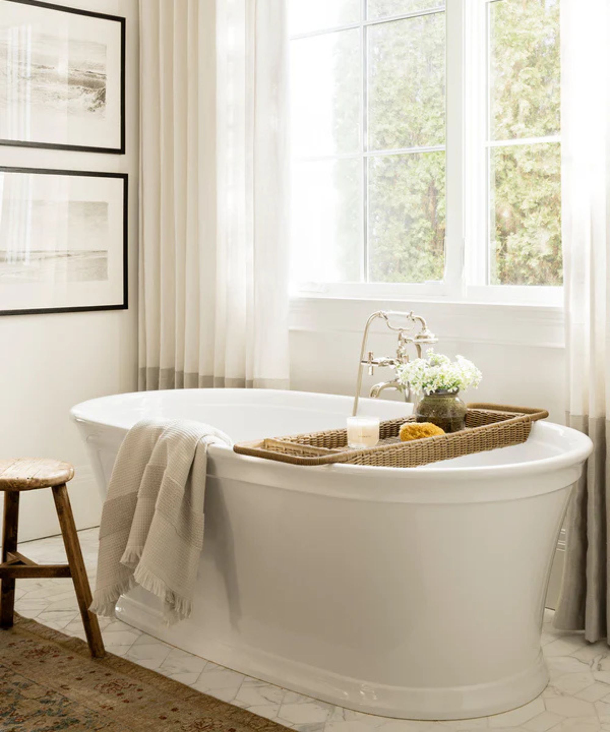 A white bathroom with a freestanding bath in front of a large window, and a rattan bath caddy displaying a candle and a small vase of flowers