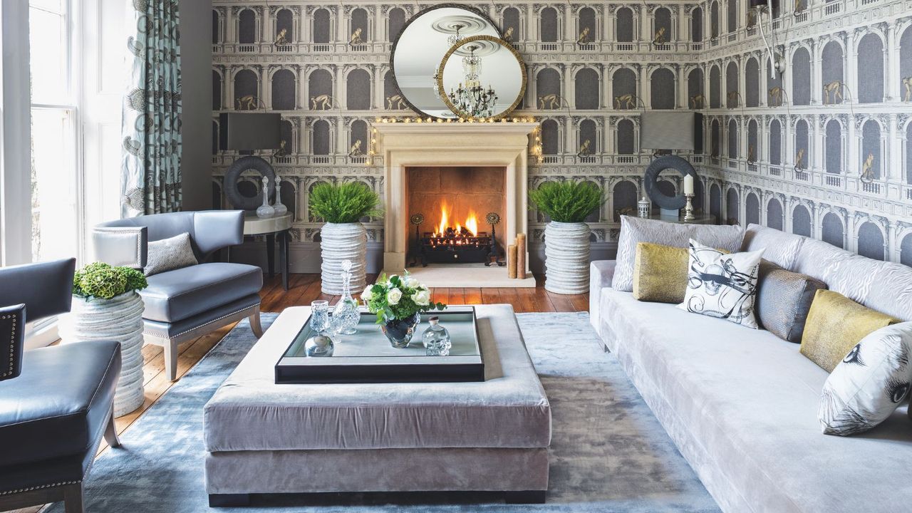 A cozy living room with statement wallpaper, a lit fire in the fireplace, a large velvet couch with matching ottoman, crushed velvet rug, and two leather chairs. On the ottoman is a tray with flowers, and above the mantelpiece are two round layered mirrors. To the left are bright, large windows with patterned curtains.