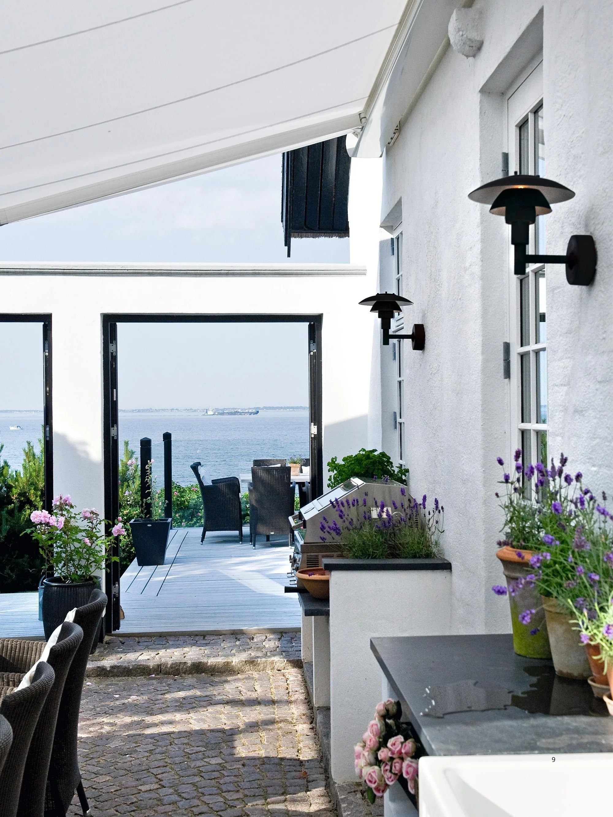 Image of a covered outdoor patio area outside of a what brick house. There is a built-in grill and lots of plant pots in view and there are two black outdoor sconce lights on the wall.