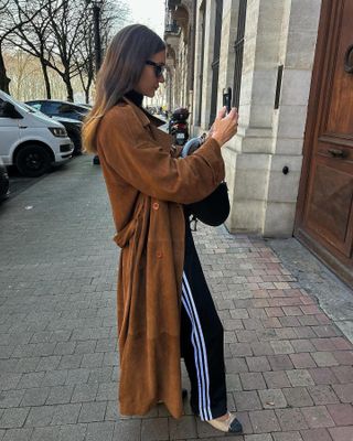 Parisian fashion influencer Anne Laure Mais posed for photos with her cell phone while wearing a long camel suede coat, Adidas sweatpants and Chanel cap-toe ballet flats.