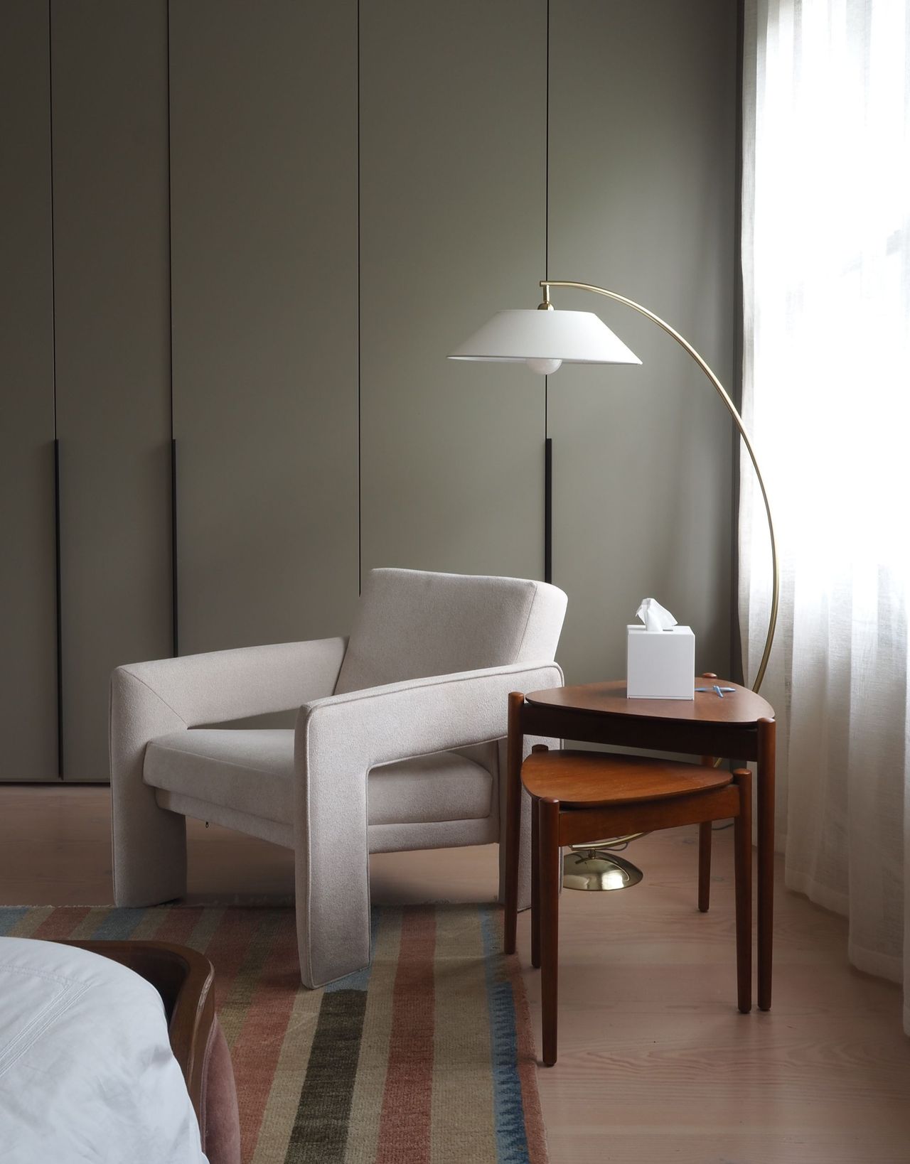 A corner seating area in a bedroom with a beige accent chair and nesting wood side tables with an arc floor lamp over the chair and sage green built-in closets behind next to sheer curtains