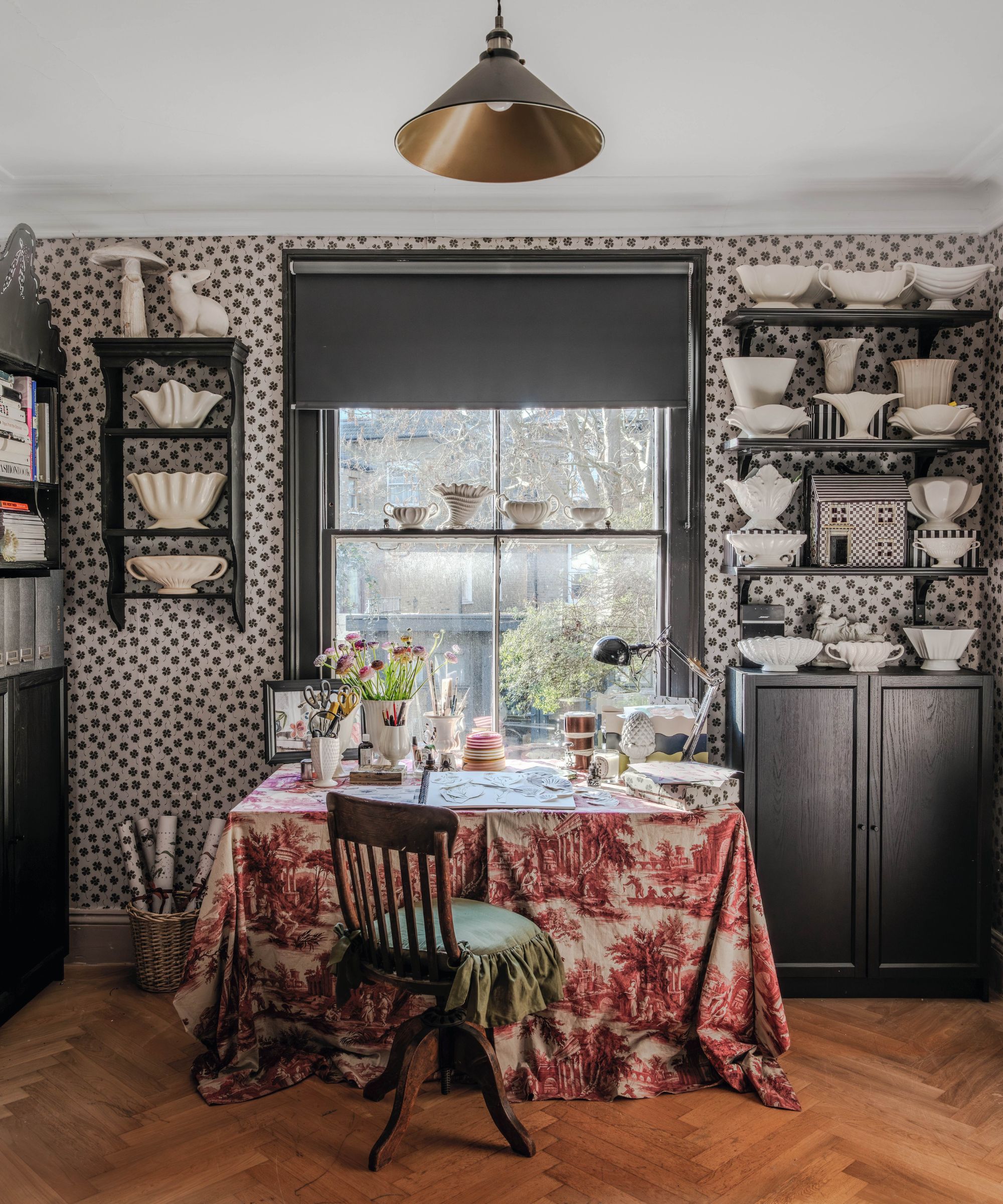 a black and white wallpapered studio room with open shelving filled with vintage ceramics and a table with a pink toile tablecloth