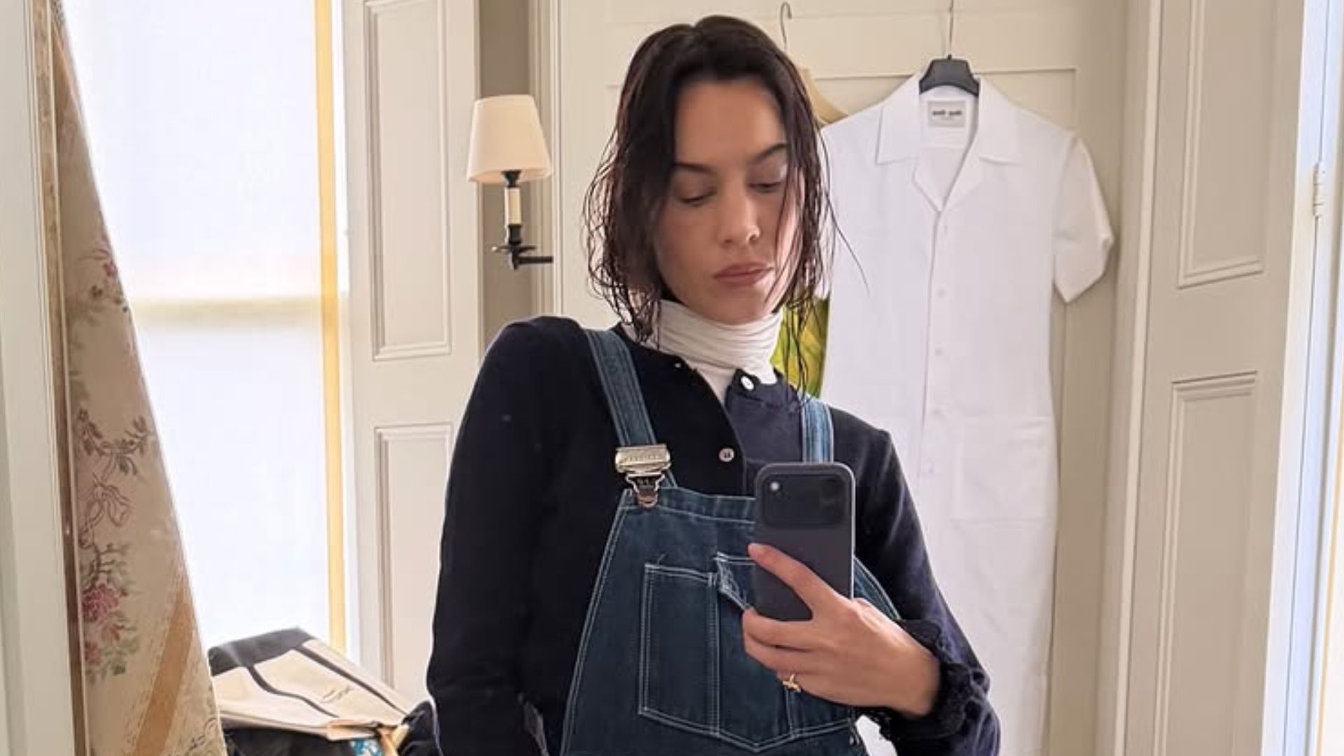 Alexa Chung takes a mirror selfie wearing dark wash dungarees with black rounded-toe boots and a white turtleneck jumper layered under a black cardigan.