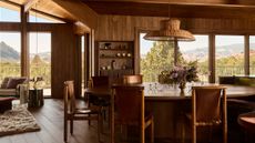 A living-dining area clad in wood with sweeping views of the Utah desert and a large dining table with leather sling seating with wood frames and a large rattan pendant light suspended above it