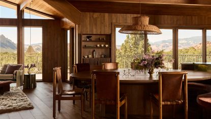 A living-dining area clad in wood with sweeping views of the Utah desert and a large dining table with leather sling seating with wood frames and a large rattan pendant light suspended above it