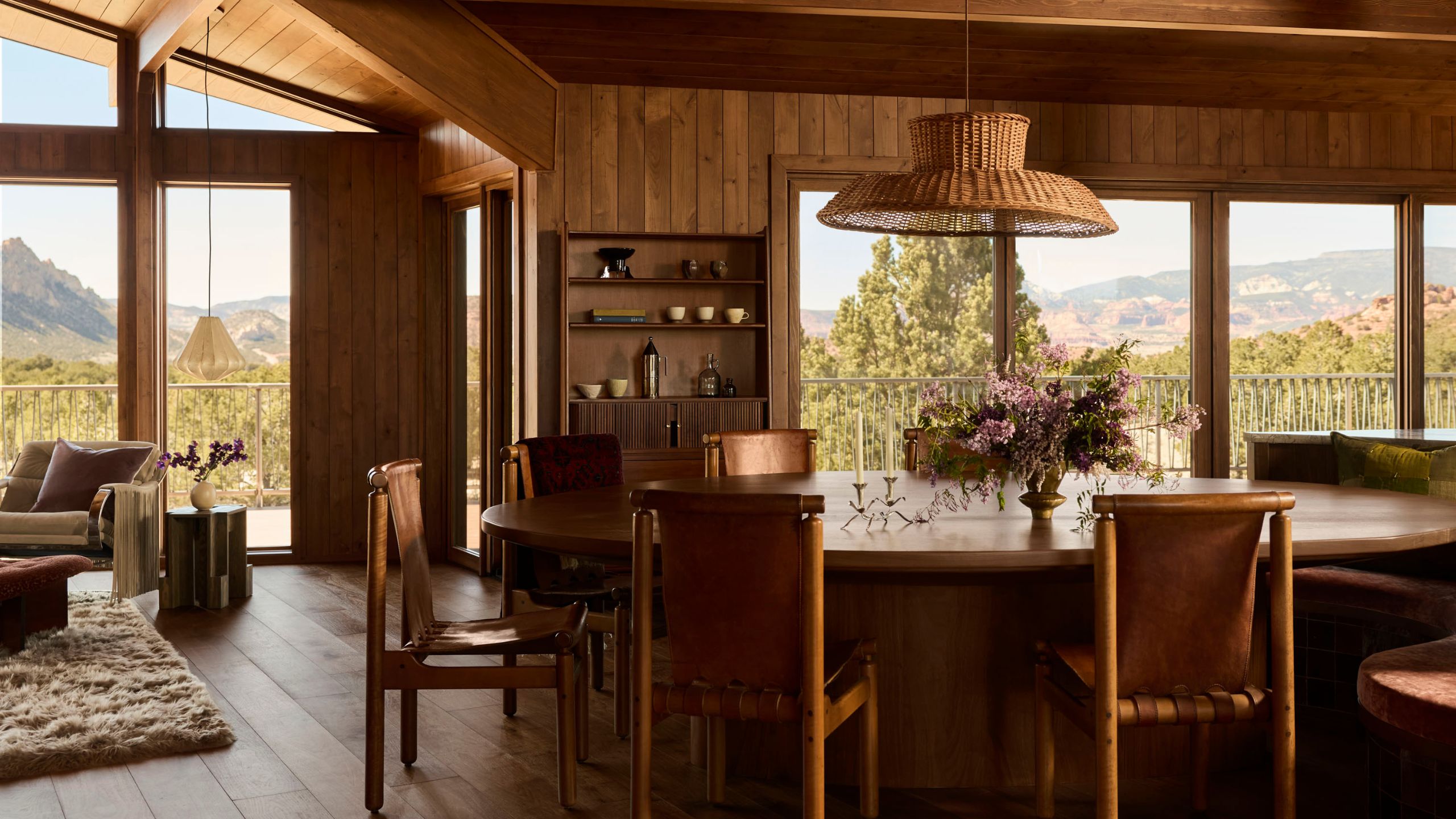A living-dining area clad in wood with sweeping views of the Utah desert and a large dining table with leather sling seating with wood frames and a large rattan pendant light suspended above it