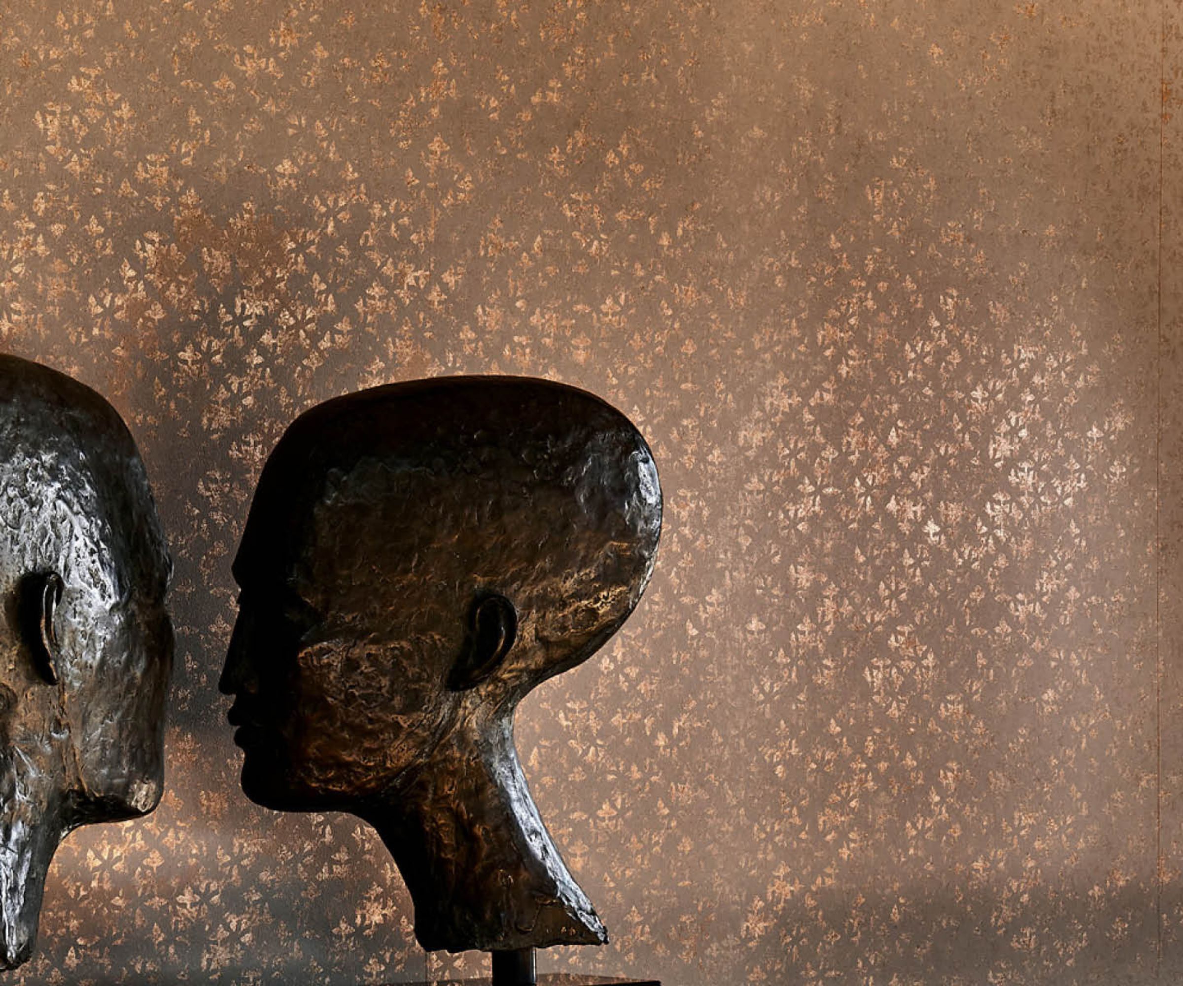 Close up of textured, shimmering beige wallpaper, two metallic head scultptures to the left of the image in front of wall