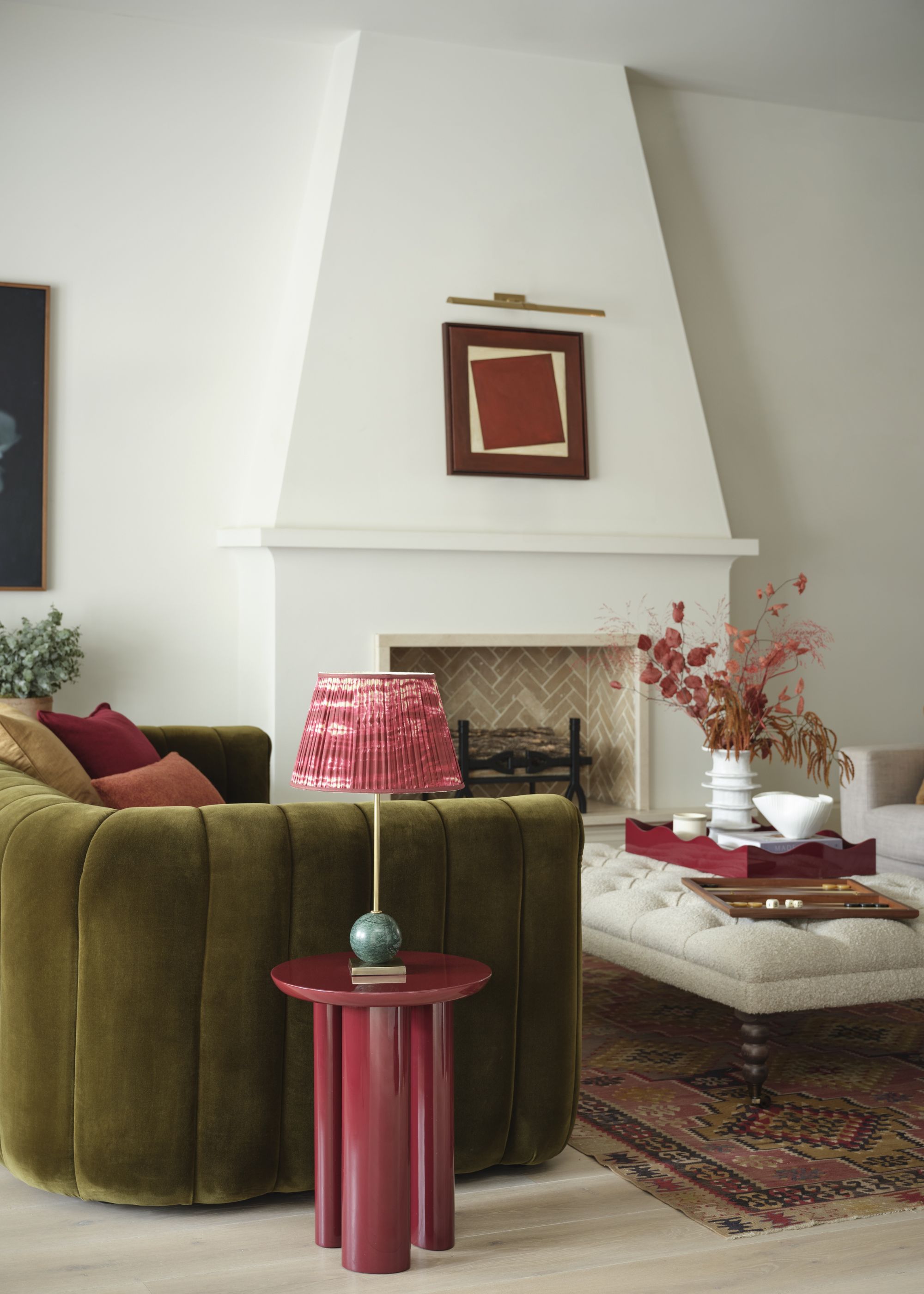 A living room with a green sofa and a pink side table with a table lamp on top. There is also an upholstered table in the middle of the living room with a curve-edges tray on top with a vase and plants