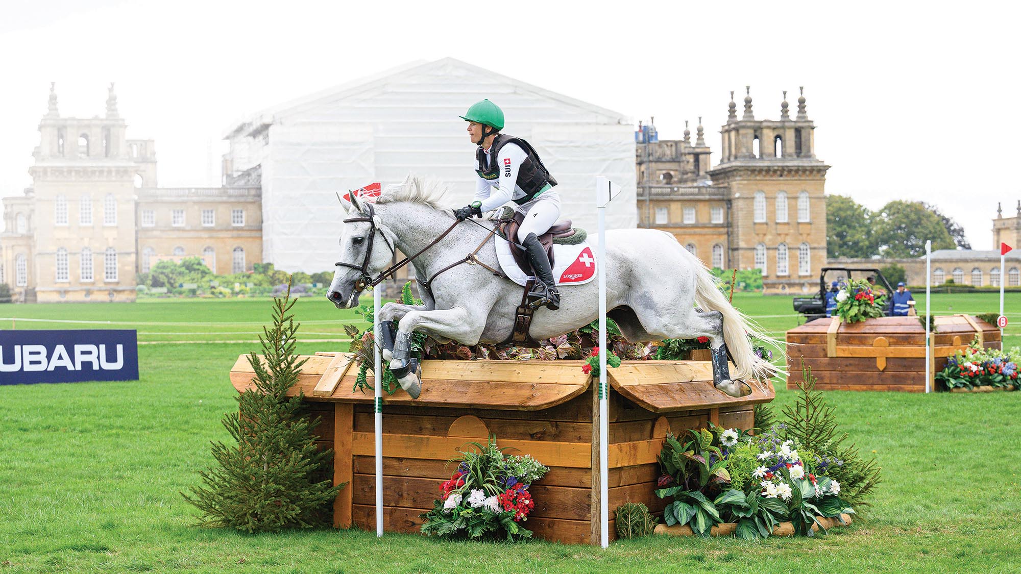 A grey horse jumps a wooden corner table in front of Blenheim Palace which is covered in scaffolding with a white cover