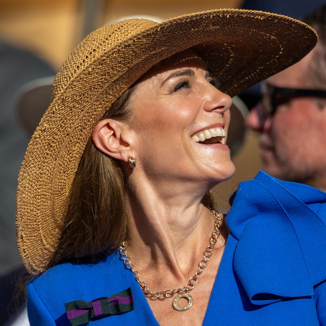 Kate Middleton wearing a blue dress and a straw hat looking up and laughing